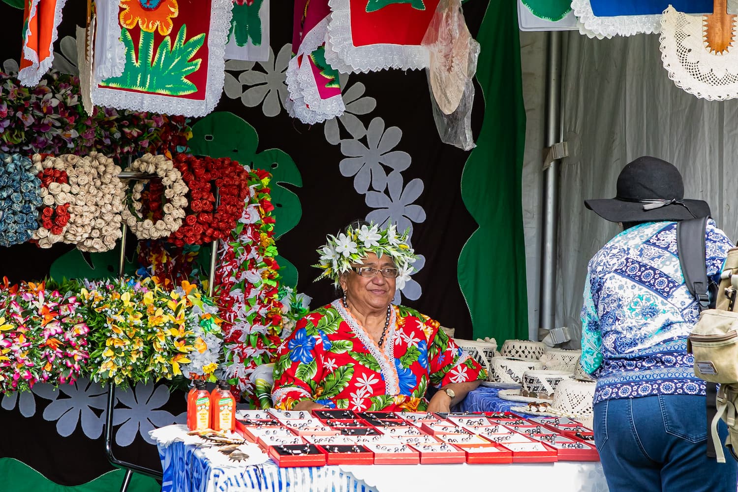 Stallholder in a colorful traditional outfit and flower crown at Auckland Pasifika Festival, surrounded by handcrafted jewelry and floral decorations.